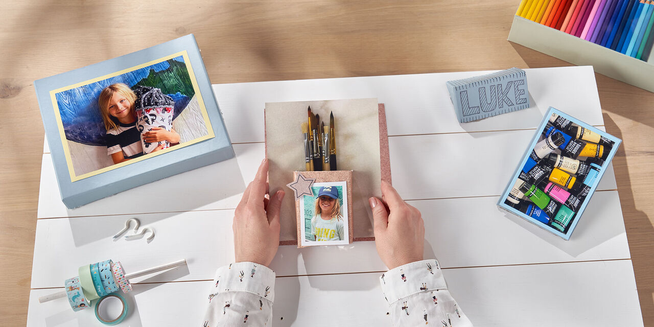 Hände halten einen selbst gebastelten Pinselhalter mit Kinderfoto über einem Brett fest. Es liegen verschiedene Bastelutensilien auf dem Tisch.